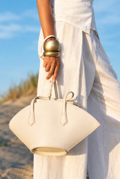 Woman in elegant white linen outfit holding beige fan shaped leather handbag at sandy beach