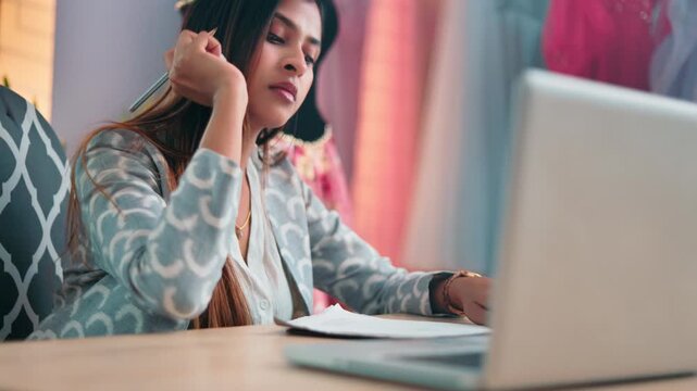 indian fashion designer drawing clothing and garment designs on a sketchbook beside a laptop in a professional atelier.