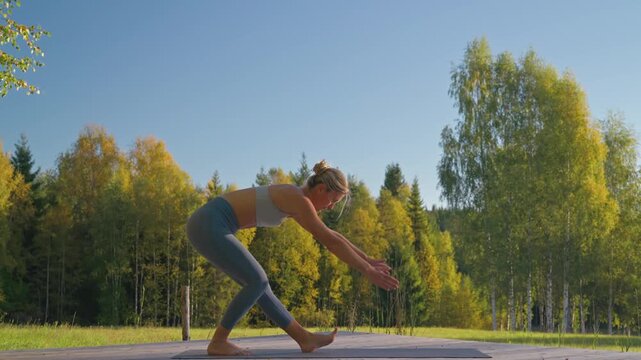Athletic Caucasian woman performing an active hamstring scoop stretch on a wooden platform in a forest, surrounded by beautiful trees, warm sunlight, and clear blue sky.