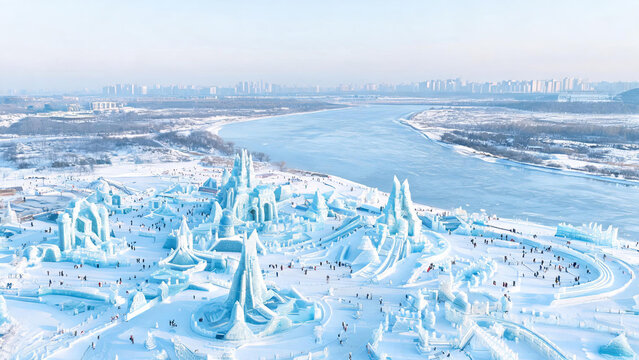 A large-scale ice sculpture festival site by a frozen river, with crowds of visitors exploring the elaborate structures on a cold winter day.