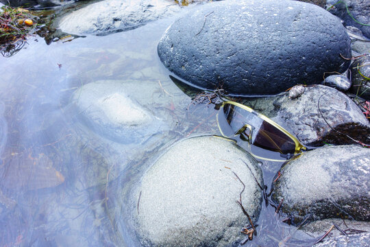 Sport sunglasses with yellow frames rest partially submerged in clear shallow water among smooth round stones at the shoreline.