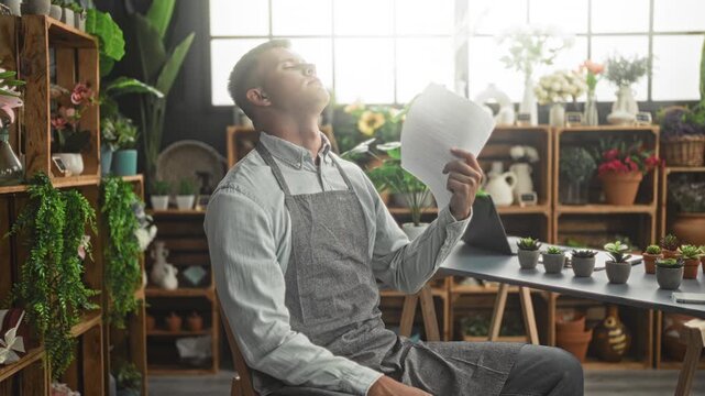 Man florist fans face with paper while seated at a flower shop counter surrounded by potted plants and shelves; fatigue.