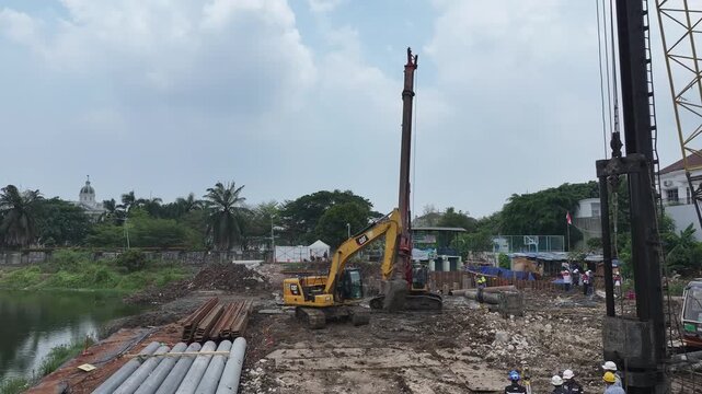 Construction site near riverbank. Workers operate heavy machinery. Red crane stands tall over muddy ground. Steel beams pile beside excavated earth. Dirt and water reflect ongoing progress