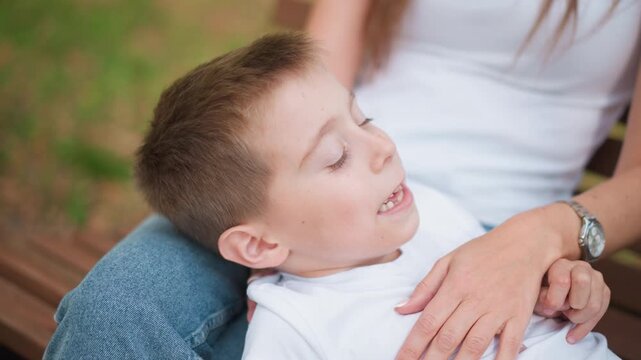 Close up of young boy resting on adult lap with boy gently supporting his head. child wears white clothing while interacting softly set against greenery and wooden bench details