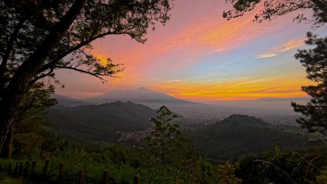 Breathtaking dusk sky over a misty mountain valley and distant peaks. Vibrant orange and pink colors frame tree silhouettes and a sleepy town nestled below majestic mountain ranges.