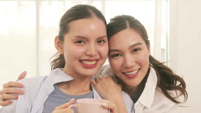 Portrait of a beautiful Asian lesbian couple relaxing at home, smiling and hugging while drinking coffee.