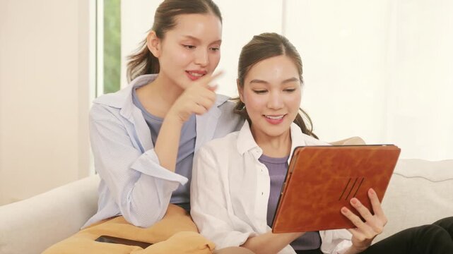 Happy young lesbian couple relaxing on a couch, browsing the internet on a tablet, and talking happily.	