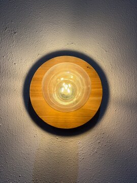 Top view of round glowing wooden wall sconce lamp with warm LED light on textured wall. Minimalist modern interior design decor light fixture providing cozy ambient lighting and home atmosphere.