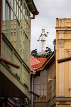 Mother of Georgia monument, Kartlis Deda, rises above traditional buildings in Tbilisi Old Town, holding sword and wine bowl while overlooking historic architecture under cloudy sky in Georgia.
