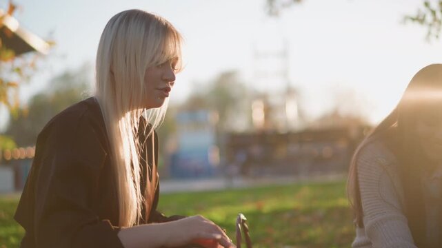 Two young women sit outdoors on grass engaged in conversation and interacting with small objects. One wears dark jacket while other has long hair visible from behind both appear relaxed