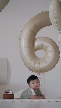 Cute Asian baby doing tummy time next to birthday cake with number 6 balloon for 6 months milestone celebration.