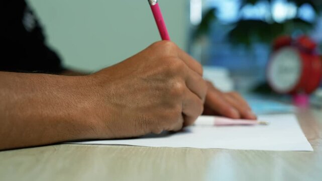 Person writing with pencil at a wooden desk, Close-up of a person writing with a pink pencil on paper at a wooden desk, with a blurred red alarm clock and soft lights in the background.