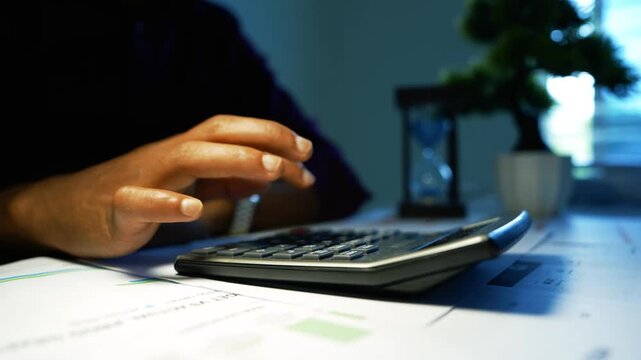 Hand Using Calculator on Office Paperwork, Close-up of a person using a calculator on paperwork at a modern office desk, with a blurred plant and window in the background.