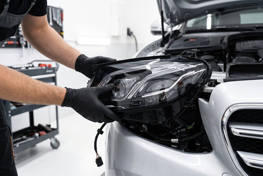 Technician hand removing headlight assembly from white vehicle in repair shop.