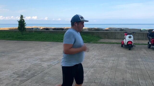 Man jogging along seaside promenade, side view with ocean and landscaped waterfront behind him, suitable for fitness, cardio training, outdoor exercise, healthy routine and active travel content