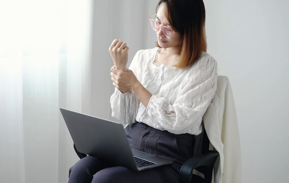 Asian businesswoman holding wrist suffering from acute pain or carpal tunnel syndrome while typing on laptop computer inside modern corporate office workspace