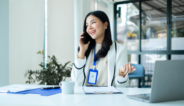 Smiling Asian businesswoman talking on smartphone at desk with laptop during client consultation in modern corporate office