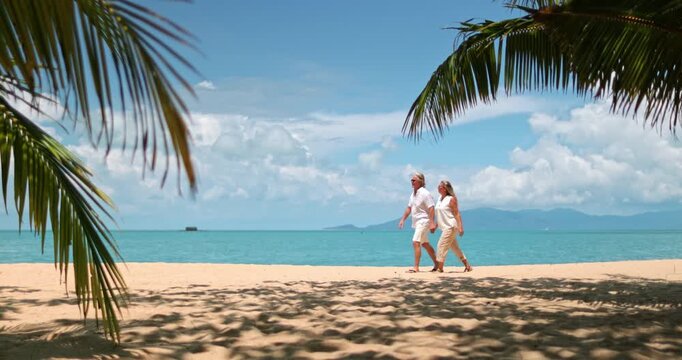 Senior couple walking hand in hand along a palm-fringed sandy beach in Samui, Thailand, enjoying a tranquil tropical vacation with a clear blue sky and calm ocean water. Romantic travel getaway