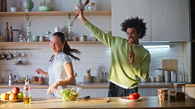 Multiethnic pair dancing home kitchen. Man singing wooden spoon cooking salad.