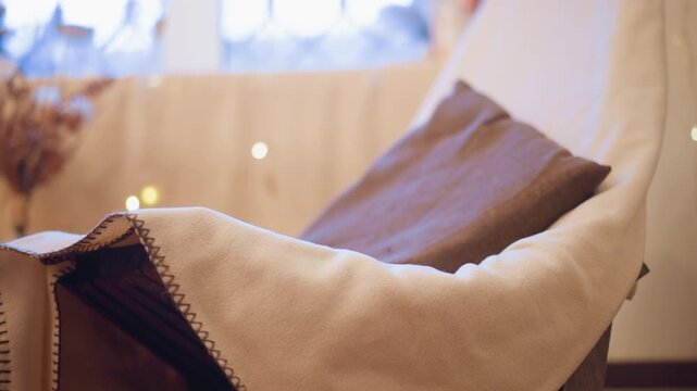 Close up of cozy white blanket with zigzag stitching draped over armchair featuring soft lighting and warm indoor ambiance. scene includes blurred decorative elements like dried flowers