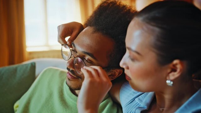 Girlfriend removes boyfriend glasses close up. Playful multiracial couple sofa.