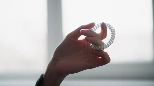 Close up of hand holding and gently rotating iridescent coiled hair tie against soft natural light. translucent plastic object reflects colors as it moves with background blurred