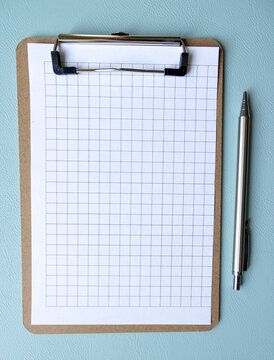 Wooden clipboard with blank grid paper and pen on solid blue backdrop