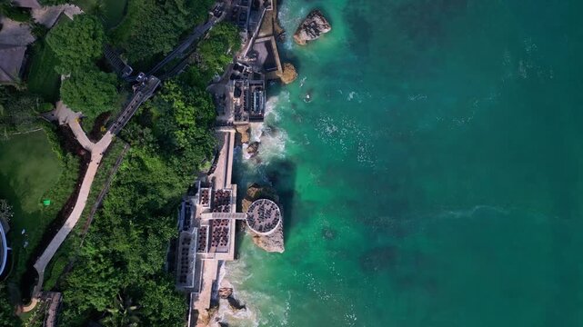 Aerial view cliff restaurant jimbaran bali, rocky coastline with turquoise water, terraced dining platforms and crashing waves, lush tropical foliage, chefs arranging tables
