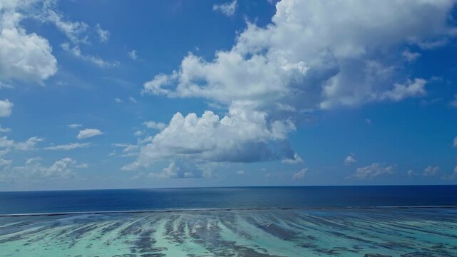 Aerial panorama shows clear turquoise water over coral reef and sandbars, opening to deep blue ocean under a sunny sky with cumulus clouds in tropical Maldives.