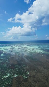 Aerial view of reef channels spreading across a turquoise lagoon into deep blue water, with sunny light and cumulus clouds forming a calm tropical Maldives seascape.