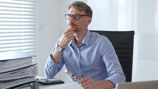 Serious male accountant is thinking while analyzing tax documents, using calculator near stacked folders, sitting in black office chair wearing crisp blue shirt. Audit and taxes in business