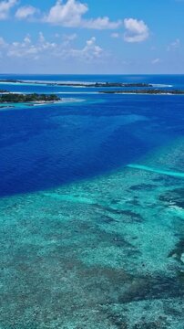 Sunlit aerial panorama of a coral reef lagoon with transparent turquoise water, layered shallows, deep blue ocean, and distant islands in a tropical holiday scene. Vertical video