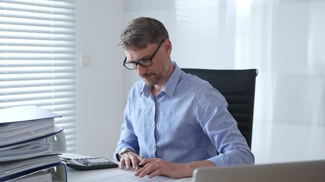 Male professional accountant wearing blue shirt and glasses is calculating invoices and managing bills while reviewing financial reports in an office setting. Audit and taxes theme in business