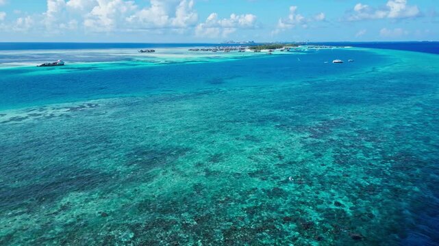 Aerial panorama shows clear turquoise water over coral reef and sandbars, opening to deep blue ocean under a sunny sky with cumulus clouds in tropical Maldives.