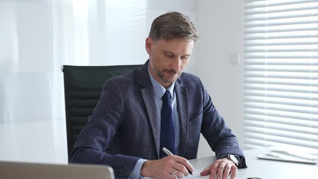 Professional businessman calculating financial data, wearing business attire while reviewing paperwork at sleek workplace desk. Audit and taxes in business