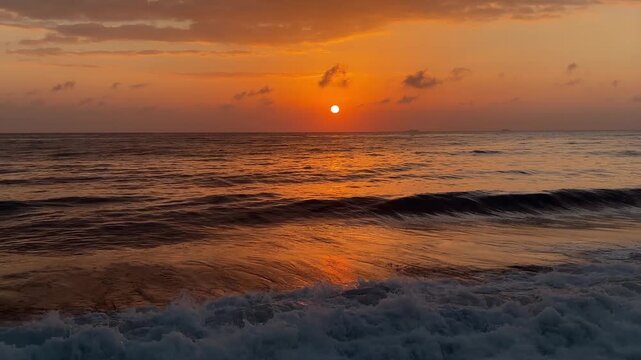 A vibrant sunset over the ocean, with waves crashing against the shore. The sky is filled with orange and yellow hues, reflecting on the water's surface.