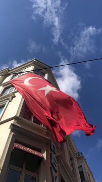 A large red flag with a white crescent and star flutters against a blue sky. The flag is prominently displayed on a building facade.