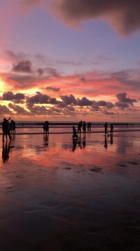 Silhouettes of people walking on a beach at sunset. The sky is filled with vibrant colors, reflecting on the wet sand. Calm ocean waves are visible.
