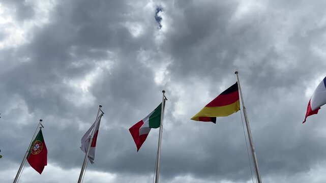 Five national flags waving against a cloudy sky. Flags include Portugal, Japan, Italy, Germany, and France. The scene conveys a sense of unity and international presence.