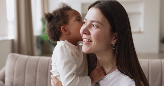 Affectionate mother sitting on sofa hugging cute biracial toddler daughter playing and talking with child enjoying sweet moment of motherhood emotional bonding playful interaction family love and care