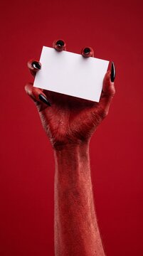A demonic hand with long black nails and red skin holds up a blank white business card, set against a vivid red background, ready to deliver a sinister message.
