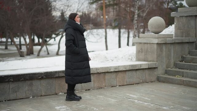 Woman in long black coat stands outdoors on snow covered ground near stone steps and spheres looking around. Woman wears boots with hands partially visible under jacket flap. background