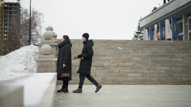 Close up of man and woman walking together in urban setting couple interacts closely as they move through public space. scene features winter weather with snow on ground surfaces stone steps