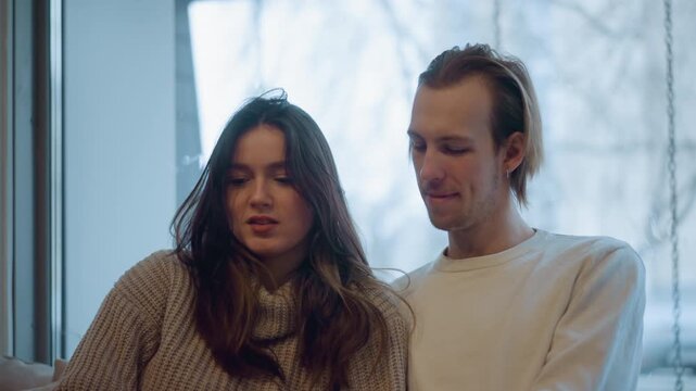 Close up of young woman and man sitting together indoors both facing forward with calm expressions. scene features soft natural light streaming through large windows behind them creating