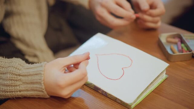 Close up of two people drawing red heart on paper with pastels hands visible. One person holds crayon while another reaches for more colors in soft indoor lighting. small metal tin