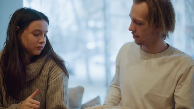 Close up of man and woman interacting with beige textured blanket indoors. Man hands over folded fabric to seated lady while they converse suggesting gift or exchange in soft lighting