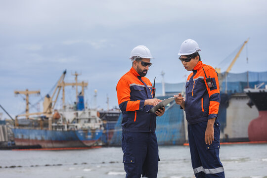engineers reviewing data on clipboard at shipyard, shipping port workers examining documentation with ships in background, maritime inspectors in safety gear discussing project operations outdoors