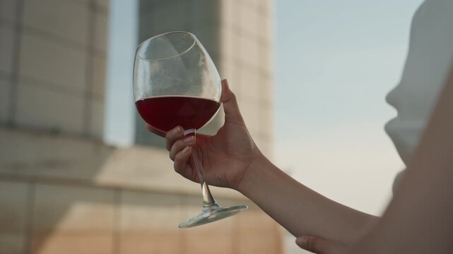 Woman holds wine glass filled with red liquid showcasing hand and partial arm. background features outdoor urban setting with architectural elements visible through soft daylight lighting