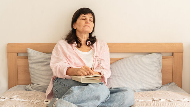 Mature woman dozing in bed with book and glasses, serene daytime rest at home, cozy and peaceful atmosphere, time for self-care. A concept of slow living, mindfulness