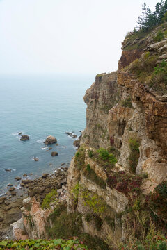 Rugged cliff rising from the sea on the northeastern China coast near Dalian, with rocky shoreline, green vegetation and calm blue water under a hazy sky